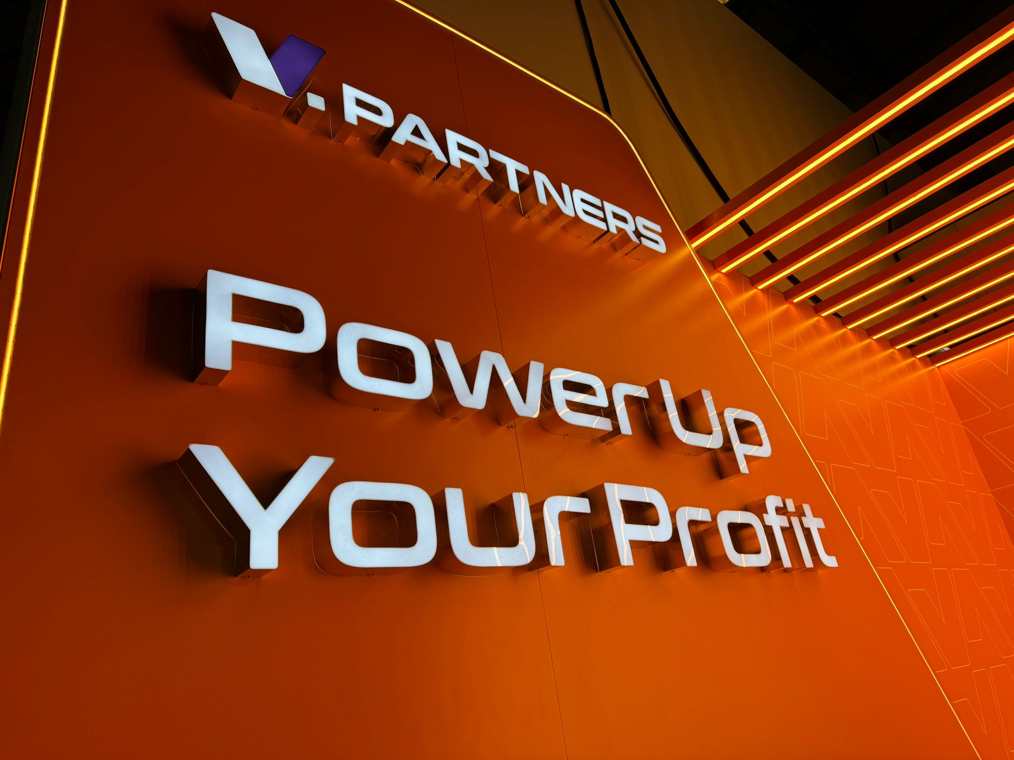 Face-lit 3D sign reading 'Power Up Your Profit' with V.Partners logo on a vivid orange wall.
